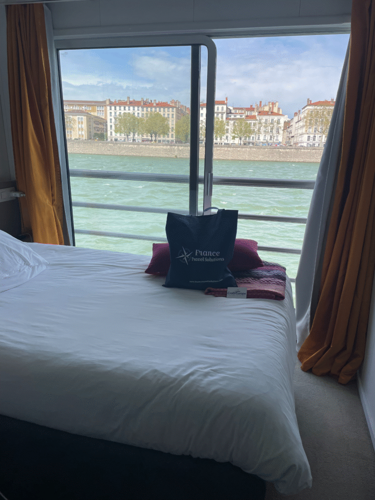 Cabin on the MS Camargue cruise ship, view of the river, and the blue France Travel Solutions tote bag on the bed